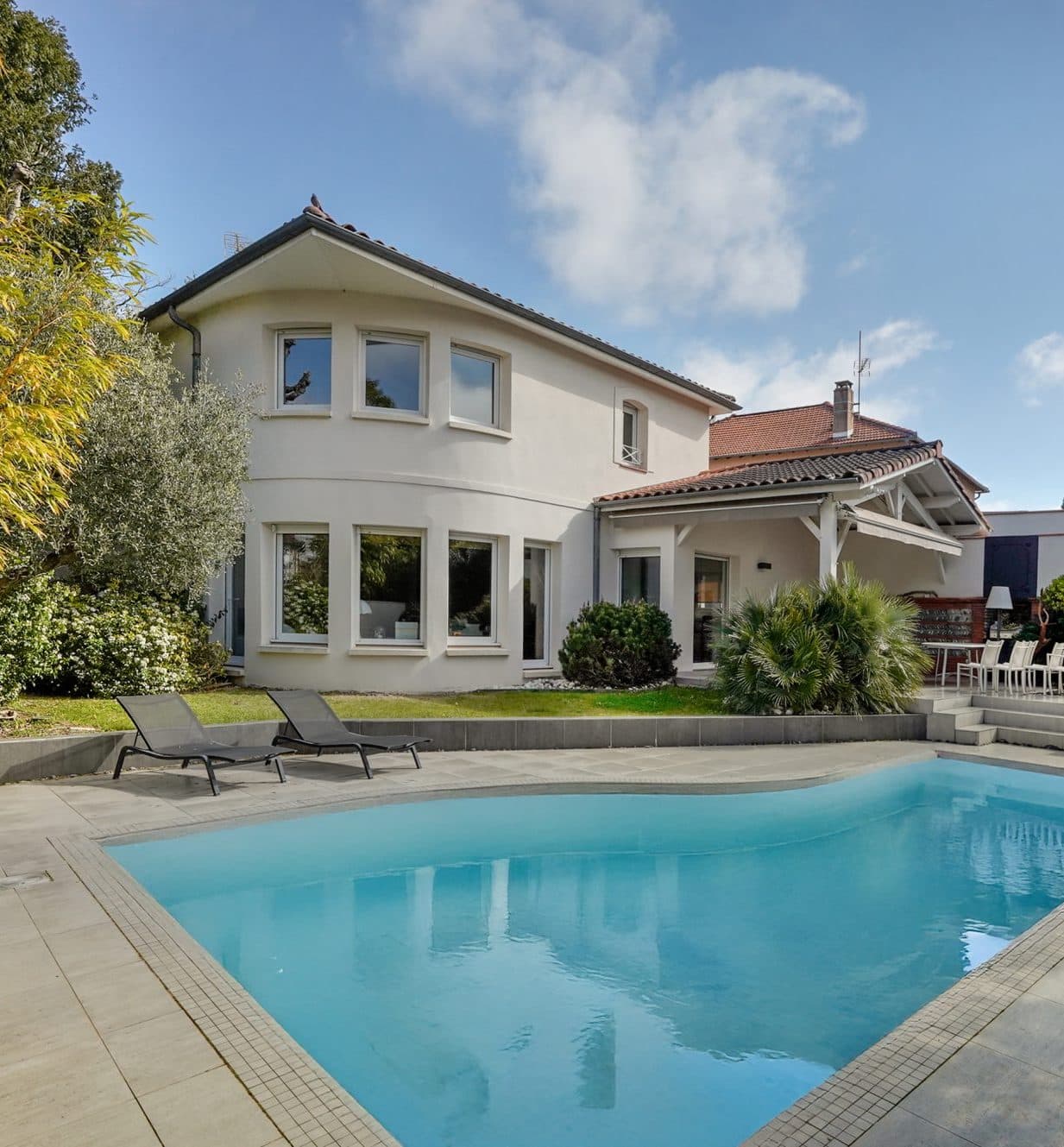 Vue extérieure d’une maison contemporaine blanche à deux étages, entourée de verdure, avec une piscine et une terrasse aménagée en premier plan.