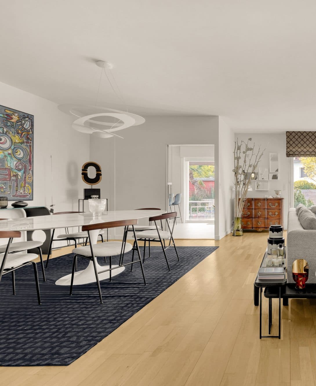 Vue intérieure d’une maison contemporaine, présentant une salle à manger épurée au design moderne, un sol en parquet clair, une table blanche entourée de chaises, un tableau d’art abstrait au mur et un vaste espace salon lumineux à l’arrière-plan.